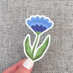 May include: A sticker featuring a stylized blue flower with a dark blue center and green leaves. The flower has a white border and is held by a hand against a gray textured background. The sticker is a floral design.
