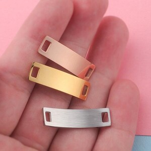 May include: A collection of rectangular metal tags in gold, rose gold, and silver. Each tag has a rectangular hole on each end. The tags are displayed against a pink and blue background.