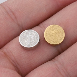 May include: Two round metal charms with a Saint Benedict medal design. One charm is silver and the other is gold.