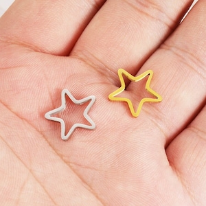 Może przedstawiać: Dwa metalowe charms w kształcie gwiazdy, jeden srebrny, a drugi złoty, pokazane są w dłoni. Charms są puste i mają prosty kształt gwiazdy.