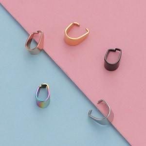 Puede incluir: Cinco baúles de colgante de metal de diferentes colores. Los baúles son de color dorado, plateado, negro, arcoíris y plateado cepillado. Los baúles están sobre un fondo rosa y azul.