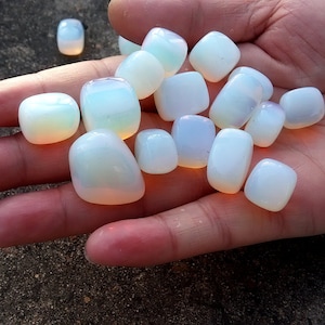 Puede incluir: Una colección de piedras lisas de ópalo blanco pulidas con destellos iridiscentes de arcoíris. Las piedras son de varios tamaños y formas, y se muestran en una mano.