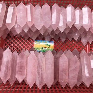 Pode incluir: Uma coleção de pontas de cristal de quartzo rosa numa cesta de plástico vermelha. Os cristais são de cor rosa suave e têm uma forma pontiaguda de dupla terminação. Os cristais estão dispostos em filas.