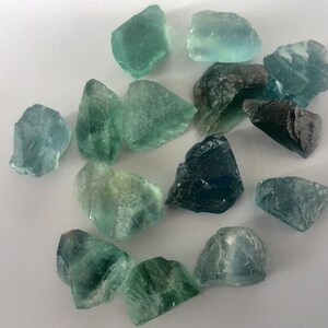 May include: A collection of green and blue fluorite crystals, some with a darker blue hue. The crystals are rough and have natural edges.
