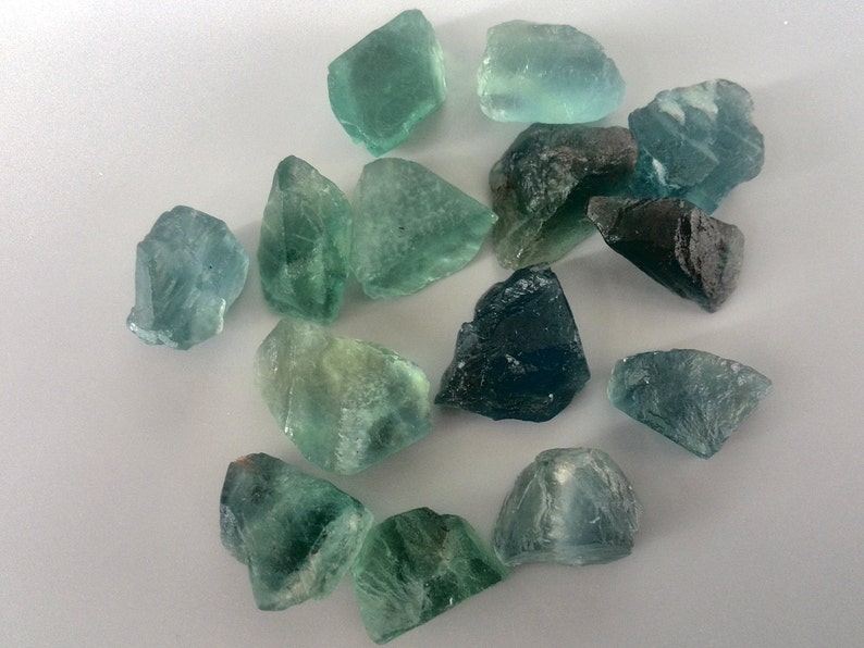 May include: A collection of green and blue fluorite crystals, some with a darker blue hue. The crystals are rough and have natural edges.