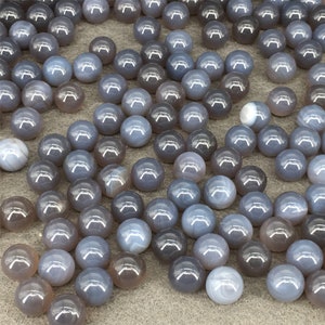 以下が含まれることがあります： グレーのアゲートビーズの集まり。それぞれが滑らかで丸い形をしており、微妙な渦巻き模様があります。ビーズは灰色の表面にランダムに配置されています。