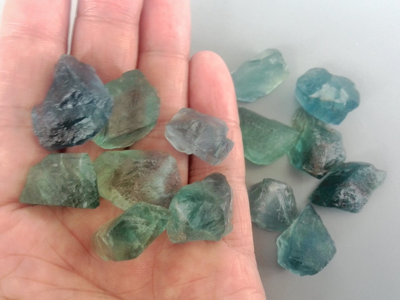 May include: A collection of green and blue fluorite crystals, some with a slight purple hue, on a hand. The crystals are various sizes and shapes, with some being more opaque than others.