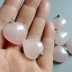 May include: Six pink heart-shaped rose quartz pendants with silver bail loops. The pendants are displayed on a hand.