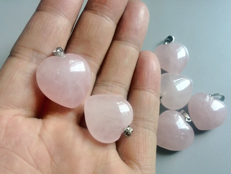 May include: Six pink heart-shaped rose quartz pendants with silver bail loops. The pendants are displayed on a hand.