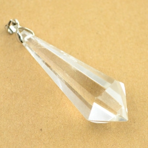 Puede incluir: Un colgante de cristal de cuarzo transparente con una anilla de plata. El cristal está facetado y tiene una punta puntiaguda.