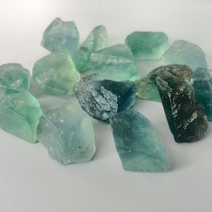 May include: A collection of teal and green fluorite crystals, some with a darker blue hue, arranged on a white surface.