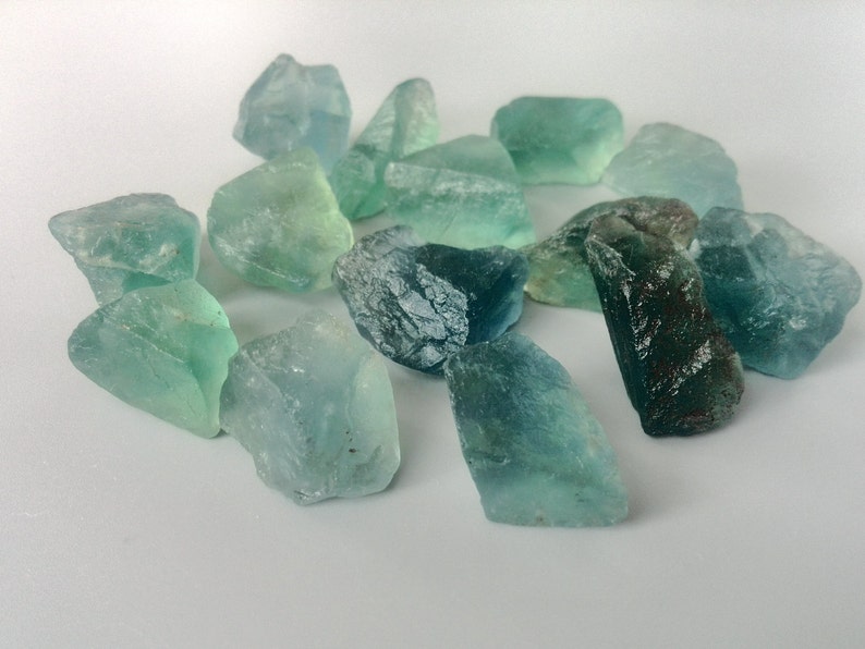 May include: A collection of teal and green fluorite crystals, some with a darker blue hue, arranged on a white surface.
