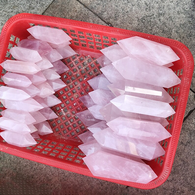 K&ouml;nnte beinhalten: Ein roter Plastikkorb gef&uuml;llt mit mehreren rosafarbenen Rosenquarz-Kristallspitzen. Die Kristalle sind doppelt terminiert und unterschiedlich gro&szlig;. Der Korb steht auf einer grauen Oberfl&auml;che.