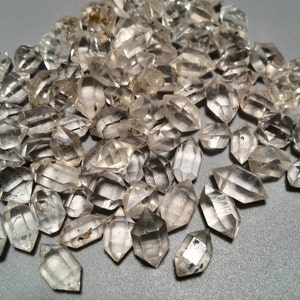 Puede incluir: Una colección de cristales de cuarzo transparentes, también conocidos como diamantes de Herkimer, con una forma de doble punta. Los cristales están dispuestos en una pila sobre una superficie gris.