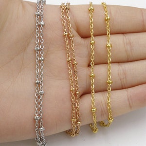Könnte beinhalten: Drei zarte Ketten mit kleinen Perlen, eine silberne, eine goldene und eine roségoldene. Die Ketten sind übereinander auf einer Hand gelegt, wodurch ihre Länge und Details zur Geltung kommen.