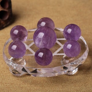 May include: A set of seven amethyst spheres arranged on a clear crystal base with a geometric design. The spheres are various shades of purple and are polished to a smooth finish.
