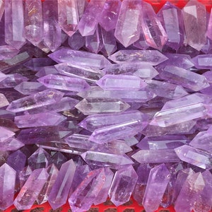 Puede incluir: Una gran colección de puntas de cristal de amatista, todas en tonos de púrpura. Las puntas están dispuestas en una cesta, algunas superpuestas. Los cristales son lisos y pulidos, con un ligero brillo.