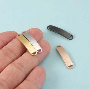 Puede incluir: Un conjunto de etiquetas de joyería metálicas en varios colores, incluyendo dorado, plateado, negro y oro rosa. Las etiquetas son rectangulares con extremos redondeados y un pequeño agujero en un extremo, para bisutería.