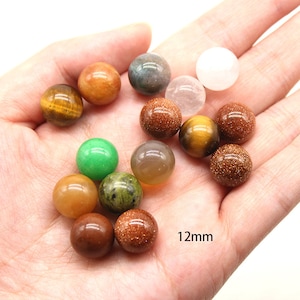 May include: A collection of 12mm round gemstone beads in various colors, including green, brown, gold, and white. The beads are displayed on a hand.