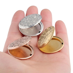 Op de afbeelding: Drie ovale medaillon hangers met ingewikkelde bloemmotieven. De hangers zijn open en laten een glad, reflecterend interieur zien. De hangers zijn zilver, goud en roségoud.