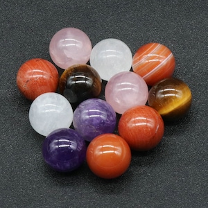 May include: A collection of 12 smooth, polished gemstone spheres in various colors, including red, orange, brown, white, pink, and purple. The spheres are arranged in a circle on a black surface.