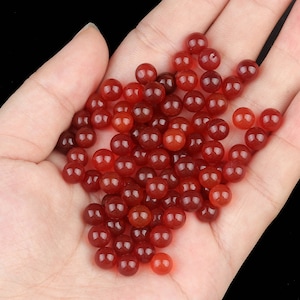 Puede incluir: Un conjunto de pequeñas cuentas redondas y pulidas en tonos rojos y anaranjados. Las cuentas son lisas y parecen translúcidas. Las cuentas están dispersas, lo que sugiere que están sueltas y listas para usar en la fabricación de joyas o manualidades.