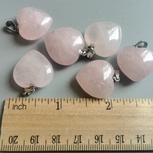 May include: Six pink heart-shaped rose quartz pendants with silver bail loops. The pendants are arranged on a wooden ruler with inch markings.