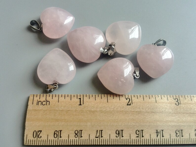 May include: Six pink heart-shaped rose quartz pendants with silver bail loops. The pendants are arranged on a wooden ruler with inch markings.