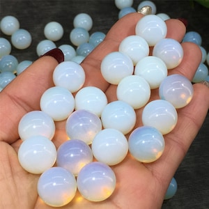 May include: A collection of smooth, white opal spheres, each with a slightly iridescent sheen. The spheres are arranged in a hand, showcasing their size and texture.