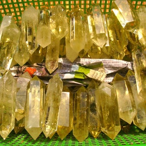 May include: A collection of golden-yellow quartz crystals, each with a pointed tip, are displayed in a green plastic basket. The crystals vary in size and shape, with a translucent appearance. The basket contains newspaper and other materials.