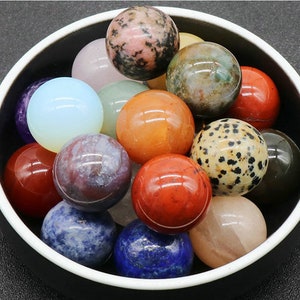 Pode incluir: Uma tigela cheia de várias esferas de pedras preciosas polidas em diferentes cores, incluindo vermelho, laranja, amarelo, verde, azul, roxo e marrom.