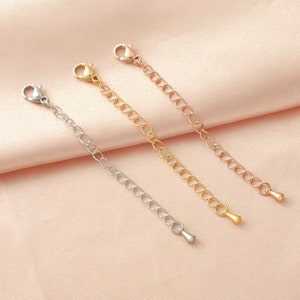 May include: Three silver chain extenders with lobster claw clasps. The extenders are different lengths and are laid out on a pink fabric background.