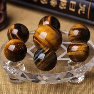 May include: Seven tiger's eye spheres arranged in a circular pattern on a clear crystal stand. The spheres are a mix of golden brown and black.