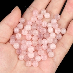 May include: A collection of small, round, rose quartz beads in varying shades of pink. The beads are smooth and polished, with a subtle translucence. The beads are held in an open hand, with a black background.