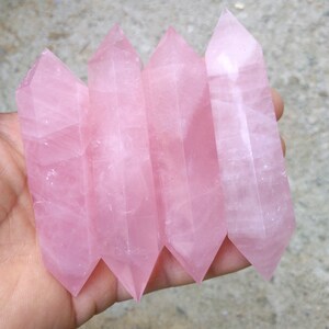 K&ouml;nnte beinhalten: Vier polierte Rosenquarz-Kristallst&auml;be. Jeder Stab hat eine zarte rosa Farbe, eine spitze Spitze und einen facettierten, rechteckigen K&ouml;rper. Die St&auml;be werden in einer Hand vor einem neutralen Hintergrund gehalten.