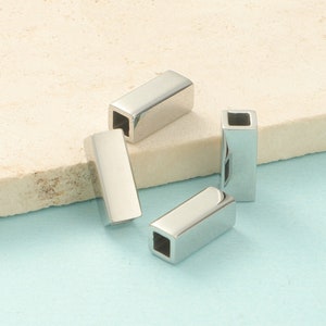 Peut inclure: Quatre entretoises rectangulaires en métal argenté avec un trou carré au centre. Les entretoises sont sur une surface beige clair.