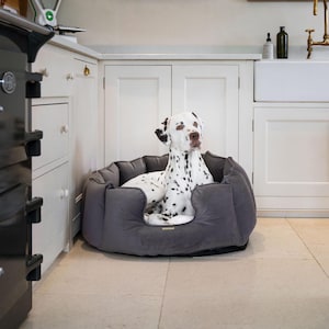 May include: A dalmatian dog relaxing in a gray dog bed with a plush, soft interior. The bed is in a kitchen setting with white cabinets and a sink.