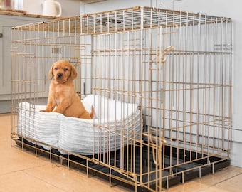 May include: A gold metal dog crate with a white and gray dog bed inside. A small, light brown puppy is sitting on the bed.