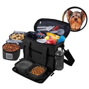 May include: Black dog travel bag with multiple compartments, including two food bowls, a water bottle holder, and a zippered pocket. The bag is filled with dog food, treats, toys, and other accessories. The bag has the text "Overland Dog Gear" on the front.