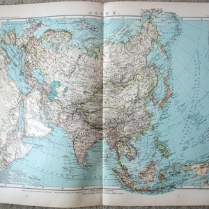 Peut inclure: Une carte ancienne de l'Asie, ouverte pour montrer le continent. Le mot "ASIEN" est en haut. Elle présente des informations géographiques détaillées, notamment les pays, les villes et les plans d'eau, dans des tons de brun, de vert et de bleu. Imprimée sur du papier vieilli.