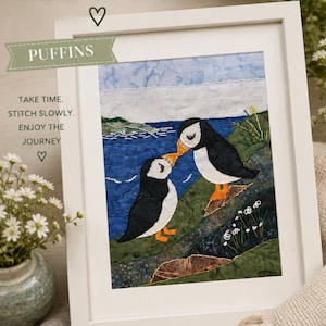 Puede incluir: Una pieza de arte textil enmarcada que muestra dos frailecillos con plumas blancas y negras y picos naranjas parados en un acantilado verde sobre un mar azul. La obra incluye el texto SLOW STITCH, PUFFINS, TAKE TIME. STITCH SLOWLY. ENJOY THE JOURNEY, STITCH, CREATE, REFLECT, CONNECT.