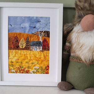 Puede incluir: Obra de arte textil enmarcada que representa un paisaje otoñal. La obra presenta una pequeña casa, árboles con colores otoñales y un campo de tonos amarillos y naranjas bajo un cielo azul. El marco es blanco.