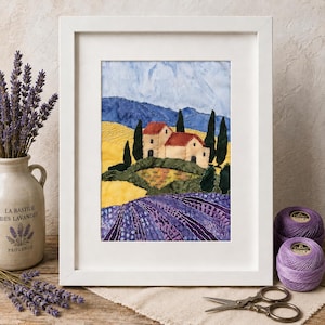 May include: A framed fabric art piece depicting a landscape with a house, yellow fields, purple lavender rows, and blue mountains. The scene features stitched details, dark green cypress trees, and a light blue sky, all encased in a white frame.