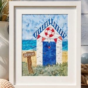 May include: A framed textile art piece featuring a white beach hut with a blue door, a red and white lifebuoy, and red bunting. The scene includes a brown sign labelled BEACH, blue ocean waves, sandy ground, and green grass, all rendered in embroidery.