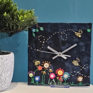 Op de afbeelding: Een zwarte stoffen klok met geborduurde bijen en bloemen. De wijzerplaat van de klok heeft een witte achtergrond met zwarte wijzers. De klok is gemonteerd op een transparante standaard.