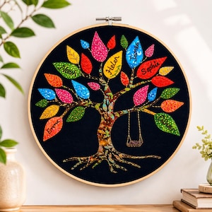 May include: A circular embroidery hoop featuring a tree with a brown trunk and roots, set against a black background. The tree has colorful patterned leaves in shades of red, yellow, blue, pink, and green. Several leaves are embroidered with the names Noah, Helen, James, Sophie, Emma, and Oliver. A small swing hangs from one of the branches.