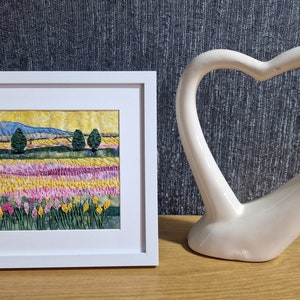 Puede incluir: Una obra de arte enmarcada que representa un paisaje con un campo de flores amarillas y rosas, dos árboles verdes y una cordillera azul en el fondo.