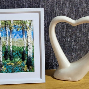 Puede incluir: Una obra de arte textil enmarcada que representa una escena de bosque con abedules y un cielo azul. Los árboles están hechos de hilo blanco y negro, y el cielo está hecho de tela azul y blanca. El fondo es una tela verde con un patrón floral.