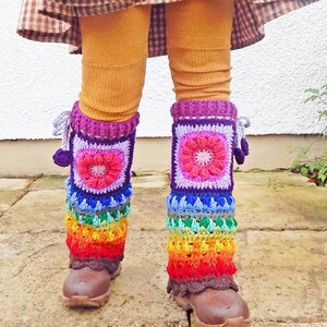 May include: Crocheted leg warmers in a rainbow design. The leg warmers feature a square design with a flower in the centre, transitioning to a vibrant spectrum of colours. They are worn over brown boots and mustard-coloured leggings.