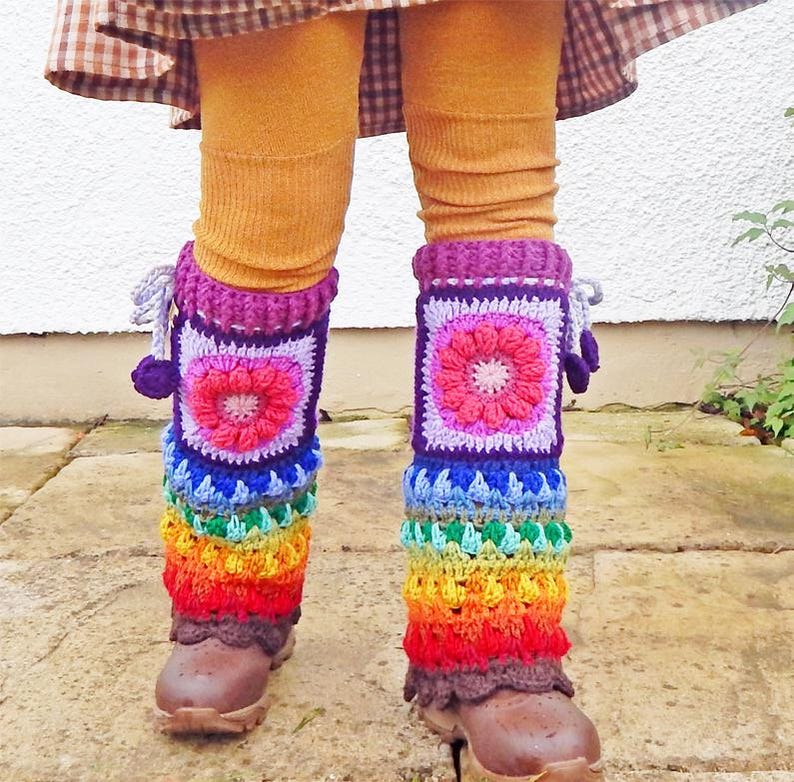 May include: Crocheted leg warmers in a rainbow design. The leg warmers feature a square design with a flower in the centre, transitioning to a vibrant spectrum of colours. They are worn over brown boots and mustard-coloured leggings.